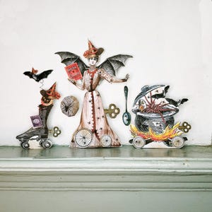 May include: A whimsical Halloween-themed display featuring paper cutouts. A witch figure with bat wings holds a book titled "Spider Cuisine." Other cutouts include a roller skate, a cauldron on wheels, a bat, and a spiderweb. The color palette includes black, red, and cream.