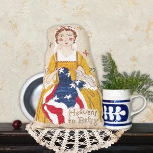 May include: A decorative, fabric doll featuring a woman in a yellow dress and a blue and red flag design. The doll has the text "Heavens to Betsy" and is displayed with a ceramic mug and a metal plate.