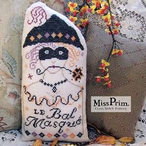 May include: A white pillow with a cross-stitch design of a masked figure with a black hat and the text "Le Bal Masque". The pillow is sitting on a brown and white patterned fabric.