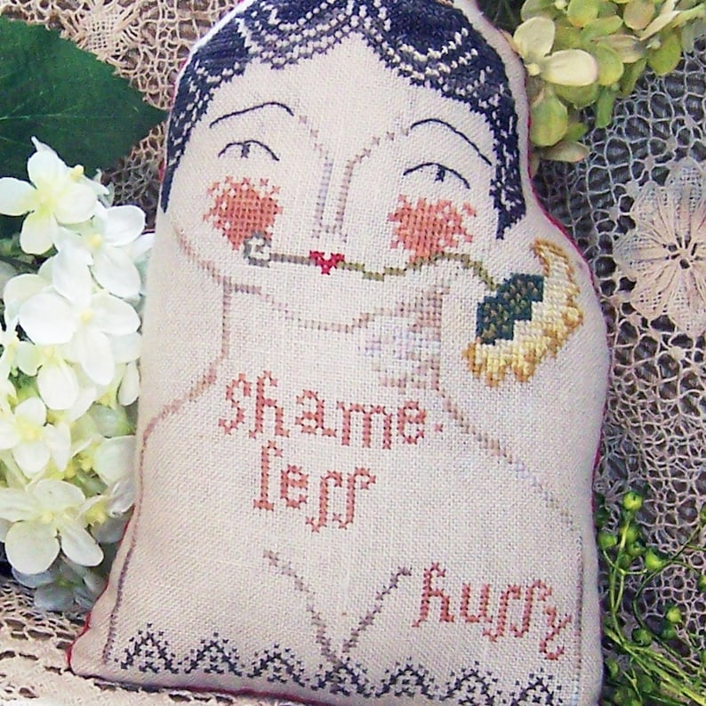 May include: A handmade, stuffed doll with a cross-stitched design. The doll features a stylized face with rosy cheeks, dark hair, and the words "shame, leff, huffy" stitched on the front. The doll is holding a flower.