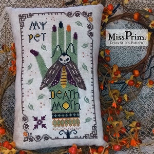 May include: Cross stitch pattern featuring a green hand with a moth on it. The hand has the words "My Pet" and "Death Moth" written on it. The pattern is framed in a brown border.