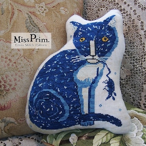 Puede incluir: Almohada de punto de cruz azul y blanco con un gato de ojos amarillos que sostiene un ratón en su boca. La almohada está hecha de lino y tiene una superficie texturizada. El texto "Miss Prim. Cross Stitch Pattern" está impreso en la almohada.