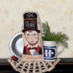 Puede incluir: Un cojín decorativo bordado con un retrato de Abraham Lincoln con sombrero de copa. El cojín tiene las palabras "Oh, Honest Abe" y "Freed" bordadas. El cojín se exhibe con un plato de metal, una taza de cerámica y cerezas.