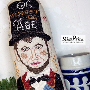 Puede incluir: Patrón de punto de cruz con un retrato de Abraham Lincoln con sombrero de copa, con el texto "Oh Honest Abe" y el año 1863. El diseño utiliza colores como negro, marrón, rojo y beige sobre tela color crema. También se muestra una taza de cerámica.