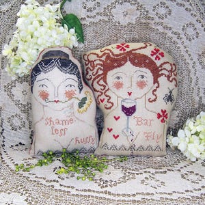 May include: Two decorative, cross-stitched pillows with female faces. One holds a flower and reads "Shame less hurry". The other holds a wine glass and reads "Bar Fly". Both have red accents and are on a lace tablecloth.