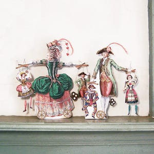 Puede incluir: Recortes de papel de figuras históricas, incluyendo una mujer con un vestido verde y rosa y un hombre con un abrigo verde. Las figuras están conectadas a palos de control de madera, lo que sugiere que son marionetas. Los recortes están en un estante.