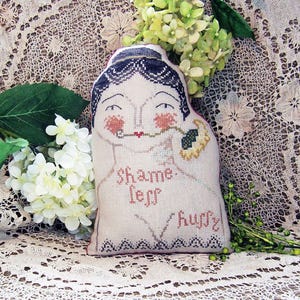 May include: A decorative, handmade pillow with a stitched design of a woman's face, featuring rosy cheeks and a red lip. The pillow has the words "shame less hugs" embroidered on it. The woman is holding a flower. The pillow is surrounded by white flowers and greenery.