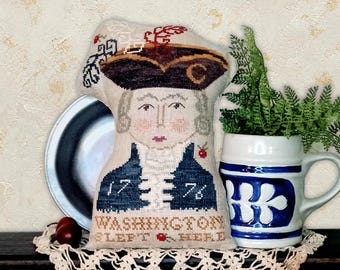May include: A decorative cushion shaped like George Washington, with a stitched design and the text "WASHINGTON SLEPT HERE" and the numbers "1776". The cushion is on a lace doily, with a ceramic mug and a metal plate in the background.