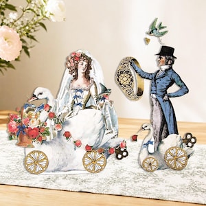 Puede incluir: Recortes de papel de estilo vintage que representan a una novia en un carruaje de cisne y a un novio con un anillo. La novia lleva un vestido blanco y un tocado floral. El novio viste un abrigo azul y un sombrero de copa. Con flores y un pájaro.