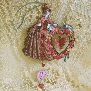 Puede incluir: Un adorno vintage para el D&iacute;a de San Valent&iacute;n con una mujer con un vestido rosa y un gran coraz&oacute;n con rosas. El adorno est&aacute; decorado con encaje y corazones.