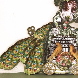 Peut inclure: Illustration vintage en papier repr&eacute;sentant une femme en robe verte avec une queue de paon. Deux oiseaux jaunes sont assis pr&egrave;s d'une cage &agrave; oiseaux sur une base &agrave; roulettes. L'illustration a une cl&eacute; &agrave; remonter et est orn&eacute;e de fleurs.