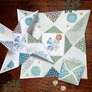 Könnte beinhalten: Ein gefalteter Papier-Wahrsager mit einem Kleeblatt-Design. Das Papier hat einen weißen Hintergrund mit blauen und grünen geometrischen Mustern und Text. Ein Cent-Stück liegt auf dem mittleren Quadrat. Der Wahrsager liegt auf einer Holzoberfläche.