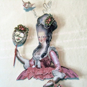 Könnte beinhalten: Eine Vintage-Papierpuppe einer Frau in einem rosa Kleid mit weißer Spitzenborte und einer großen weißen Perücke. Sie hält einen roten Stock mit einem herzförmigen Spiegel, auf dem "Amour" steht. Die Puppe ist von einem weißen Hintergrund umgeben.