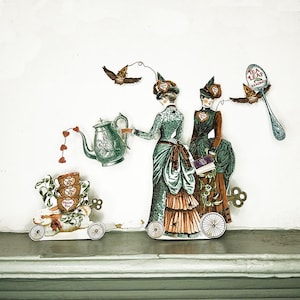 Peut inclure: D&eacute;coupages de papier fantaisistes repr&eacute;sentant des femmes de l'&eacute;poque victorienne, une th&eacute;i&egrave;re, un lapin sur roues et un hibou avec une cuill&egrave;re. Les femmes portent des robes &eacute;labor&eacute;es. La th&eacute;i&egrave;re a un jet en forme de c&oelig;ur. Le hibou tient une cuill&egrave;re avec "Tea Leaf Reading".