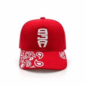 May include: A bright red baseball cap featuring a white embroidered logo on the front panel. The curved brim is decorated with a white paisley pattern that extends across the front, providing a distinct contrast against the solid red fabric.
