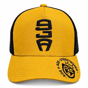 Könnte beinhalten: Eine senfgelbe Baseballkappe mit schwarzem Netzrücken. Die Vorderseite zeigt ein vertikal gesticktes schwarzes Logo mit der Aufschrift ALD. Der Schirm zeigt eine schwarze kreisförmige Grafik mit dem Text ADE + ADE STREETWEAR und einem Gesichtssymbol.