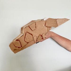 Puede incluir: Escultura de cartón marrón de una cabeza de dinosaurio con detalles rojos. La cabeza es sostenida por un brazo. La escultura tiene formas geométricas recortadas en el cartón.