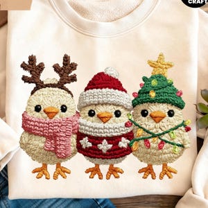Könnte beinhalten: Cremefarbenes Sweatshirt mit drei gestickten Küken in festlicher Weihnachtskleidung. Ein Küken trägt Rentiergeweih und einen rosa Schal, ein anderes einen Weihnachtsmannhut und -pullover, und das letzte einen Weihnachtsbaumhut mit Lichtern.