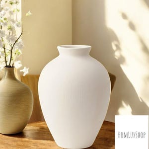 Könnte beinhalten: Eine weiße Keramikvase mit runder, bauchiger Form. Die Vase steht auf einer Holzoberfläche.