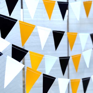 May include: A string of triangular pennant flags in black, white, and yellow. The flags are strung together on a white string, creating a festive and decorative banner. The pennants are made of fabric and are suitable for parties or celebrations.