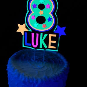 Puede incluir: Un adorno para pastel de color verde ne&oacute;n con el n&uacute;mero ocho, estrellas de colores y el nombre "LUKE" en colores ne&oacute;n. El adorno est&aacute; sobre un pastel blanco glaseado con glaseado azul.
