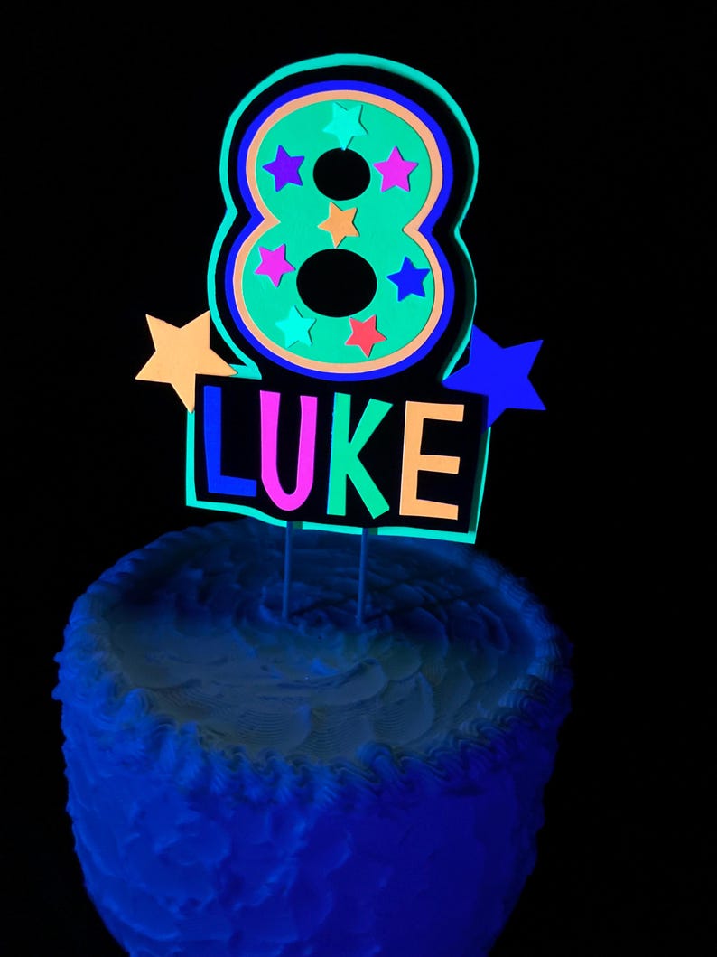 Puede incluir: Un adorno para pastel de color verde ne&oacute;n con el n&uacute;mero ocho, estrellas de colores y el nombre "LUKE" en colores ne&oacute;n. El adorno est&aacute; sobre un pastel blanco glaseado con glaseado azul.
