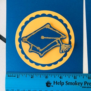 May include: A blue and yellow graduation banner with a gold glitter graduation cap in the center. The banner is decorated with a scalloped circle around the cap. The banner is 6 inches long.