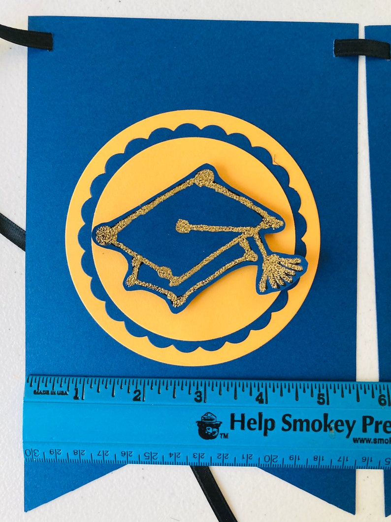 May include: A blue and yellow graduation banner with a gold glitter graduation cap in the center. The banner is decorated with a scalloped circle around the cap. The banner is 6 inches long.