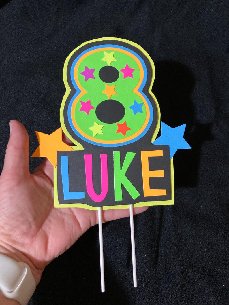 Puede incluir: Un adorno para pastel de color verde ne&oacute;n y negro con el n&uacute;mero 8 y el nombre "LUKE" en letras de colores. El n&uacute;mero 8 est&aacute; rodeado de estrellas de colores. El adorno est&aacute; en dos palos blancos.