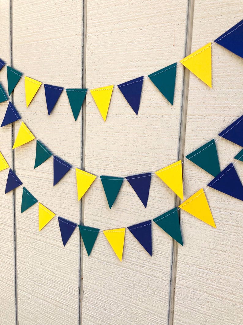 Peut inclure: Une guirlande festive compos&eacute;e de fanions triangulaires jaunes, bleu marine et vert sarcelle. Les fanions sont reli&eacute;s par un fil blanc, cr&eacute;ant une guirlande d&eacute;corative. La guirlande est affich&eacute;e sur un fond en bois blanc.