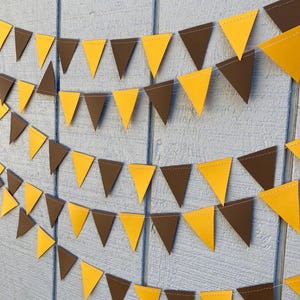 May include: A festive banner of alternating yellow and brown triangle flags strung on white thread. The pennants are arranged in multiple rows against a light blue wooden background, creating a decorative display.