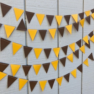 May include: A decorative banner with alternating brown and yellow triangular flags. The pennants are strung together and hang against a light blue wooden background. The banner is suitable for parties and celebrations.