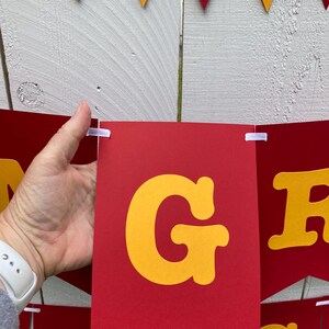 May include: Red and yellow pennant banner with the word "GRAD" in large, yellow, block letters. The banner is strung together with white string.