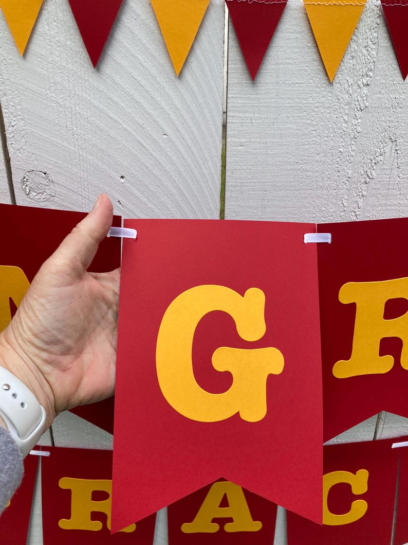 May include: Red and yellow pennant banner with the word "GRAD" in large, yellow, block letters. The banner is strung together with white string.