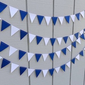 May include: Several strands of triangular pennant flags in alternating white and royal blue. The flags are strung together and hang against a light gray background, creating a festive and decorative display. Perfect for parties and celebrations.