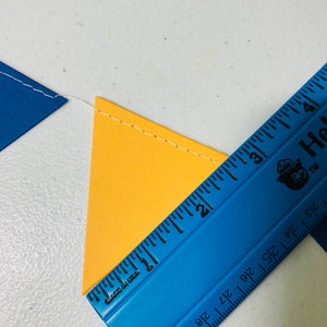 May include: A blue ruler with black markings and the text "Made in USA" and "Hallmark" is placed on top of a yellow triangle. The ruler is measuring the triangle, which is 2 inches wide.