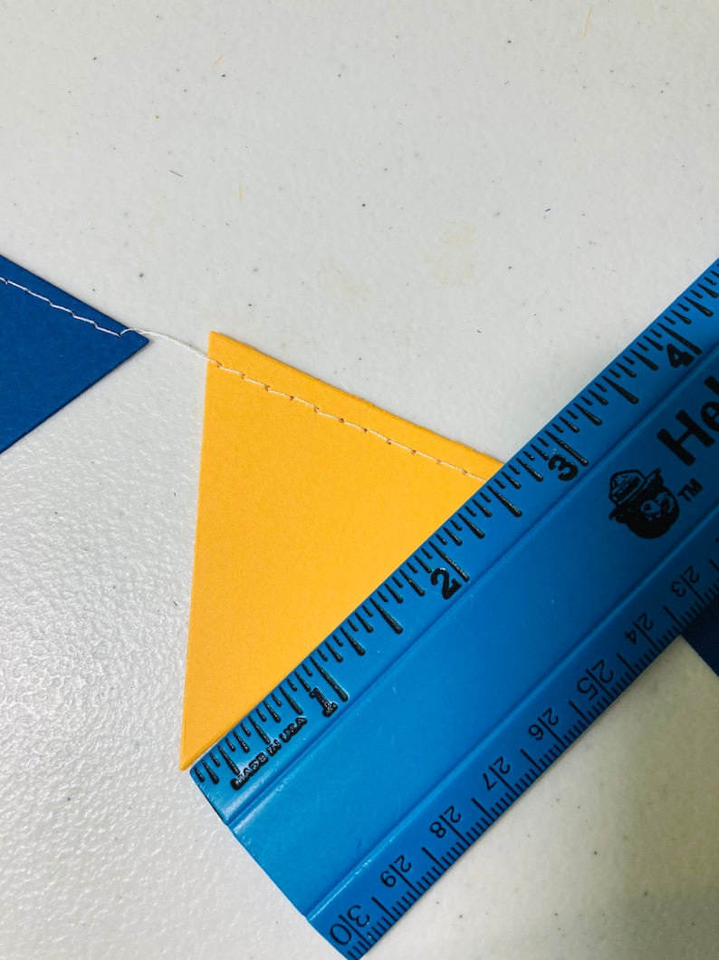 May include: A blue ruler with black markings and the text "Made in USA" and "Hallmark" is placed on top of a yellow triangle. The ruler is measuring the triangle, which is 2 inches wide.