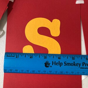 May include: Red banner with a yellow letter "S" cut out. The banner is approximately 6 inches wide. The banner has white ribbon attached to the top corners.