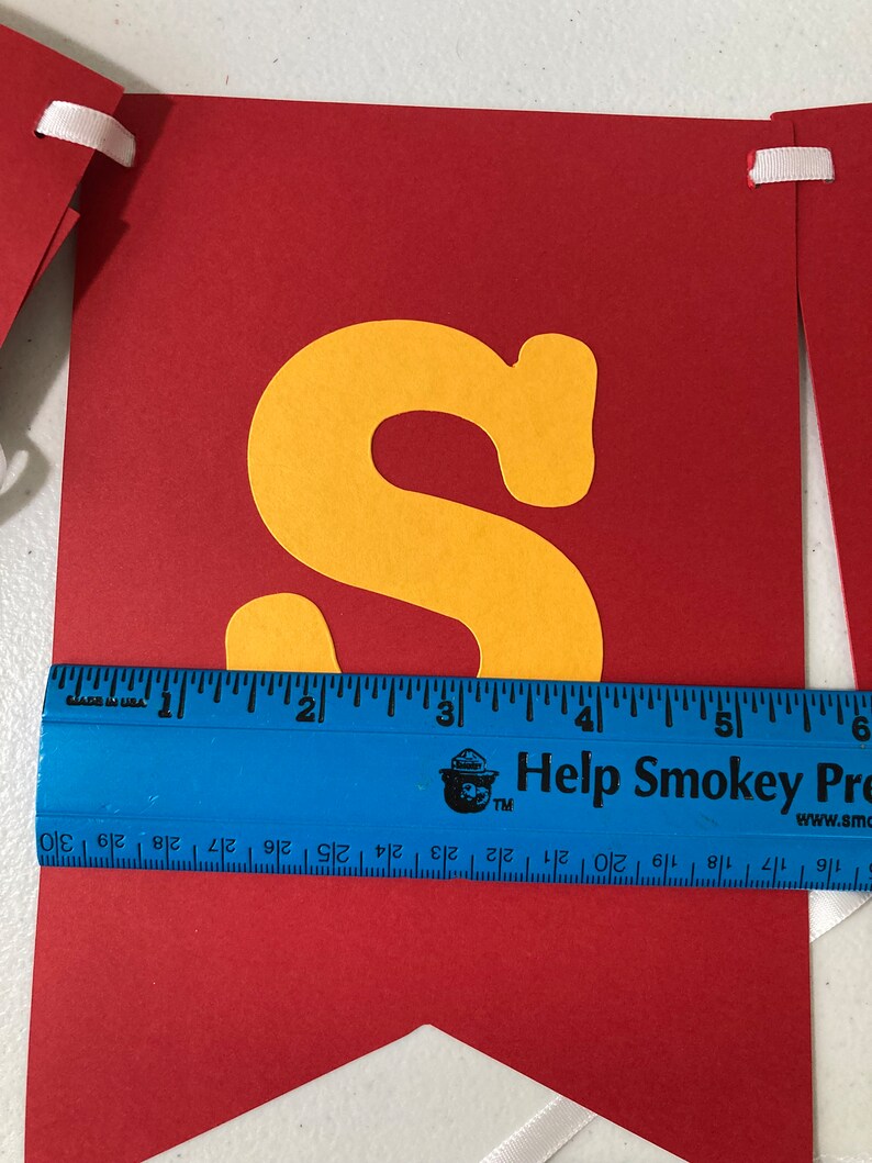 May include: Red banner with a yellow letter "S" cut out. The banner is approximately 6 inches wide. The banner has white ribbon attached to the top corners.