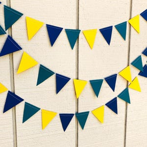 Peut inclure: Une guirlande festive compos&eacute;e de fanions triangulaires jaunes, bleu sarcelle et bleu marine. Les fanions sont enfil&eacute;s sur un fil blanc, cr&eacute;ant une guirlande joyeuse et d&eacute;corative. Le fond est une surface en bois blanc.