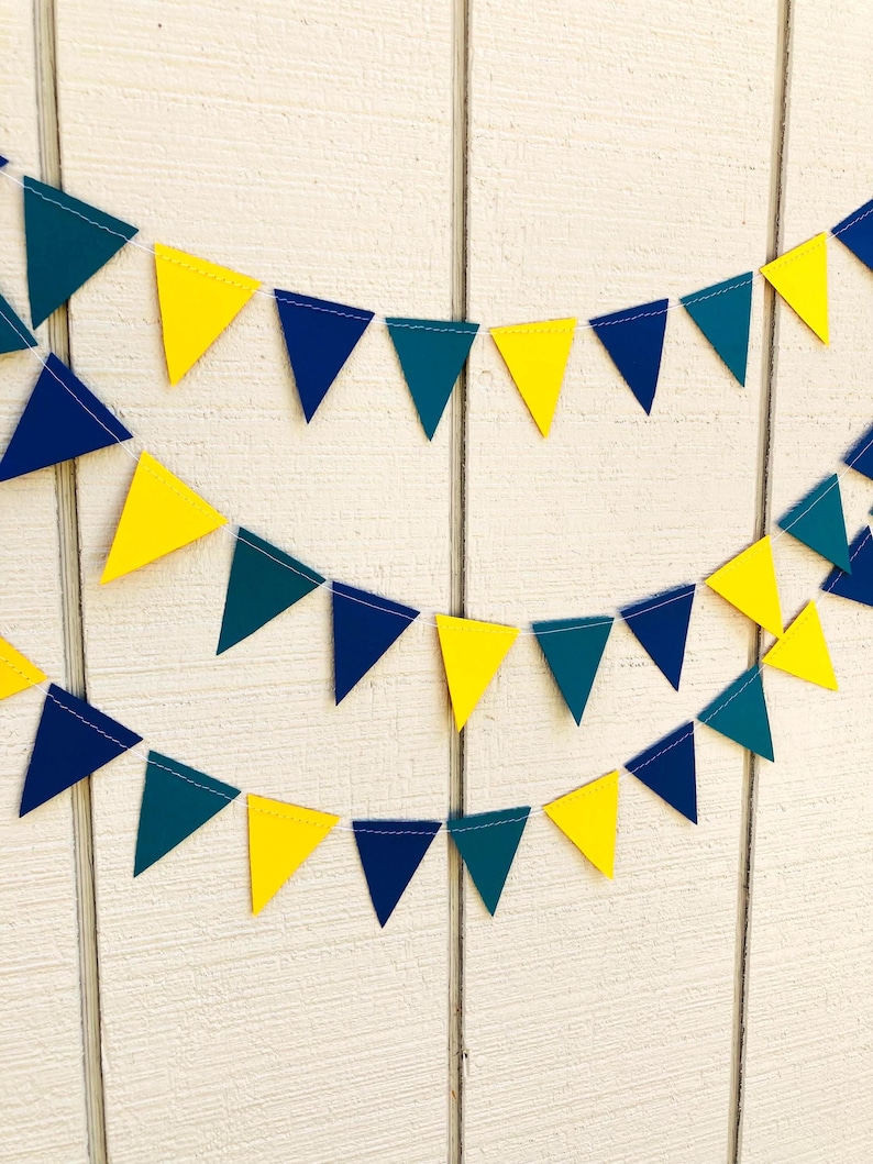 Peut inclure: Une guirlande festive compos&eacute;e de fanions triangulaires jaunes, bleu sarcelle et bleu marine. Les fanions sont enfil&eacute;s sur un fil blanc, cr&eacute;ant une guirlande joyeuse et d&eacute;corative. Le fond est une surface en bois blanc.