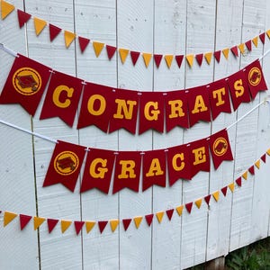 May include: Red and gold graduation banner with the words "Congrats Grace" and graduation caps. The banner is strung on a white string and is hanging on a white wooden fence.