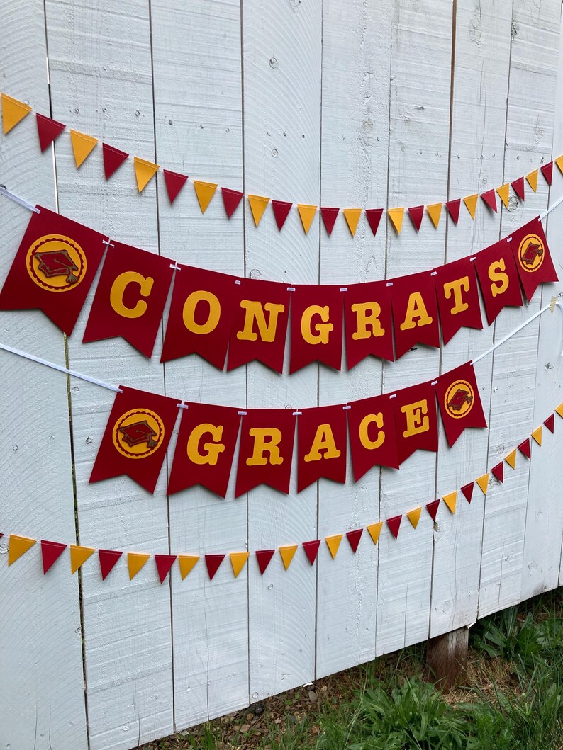 May include: Red and gold graduation banner with the words "Congrats Grace" and graduation caps. The banner is strung on a white string and is hanging on a white wooden fence.