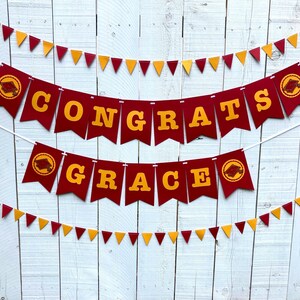 May include: Red and gold graduation banner with the words "Congrats Grace" on it. The banner is strung up on a white wooden fence.