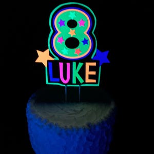 Puede incluir: Un adorno para pastel de color verde ne&oacute;n con el n&uacute;mero 8, estrellas de colores y el nombre "LUKE" en colores ne&oacute;n. El adorno para pastel est&aacute; sobre un pastel blanco glaseado.