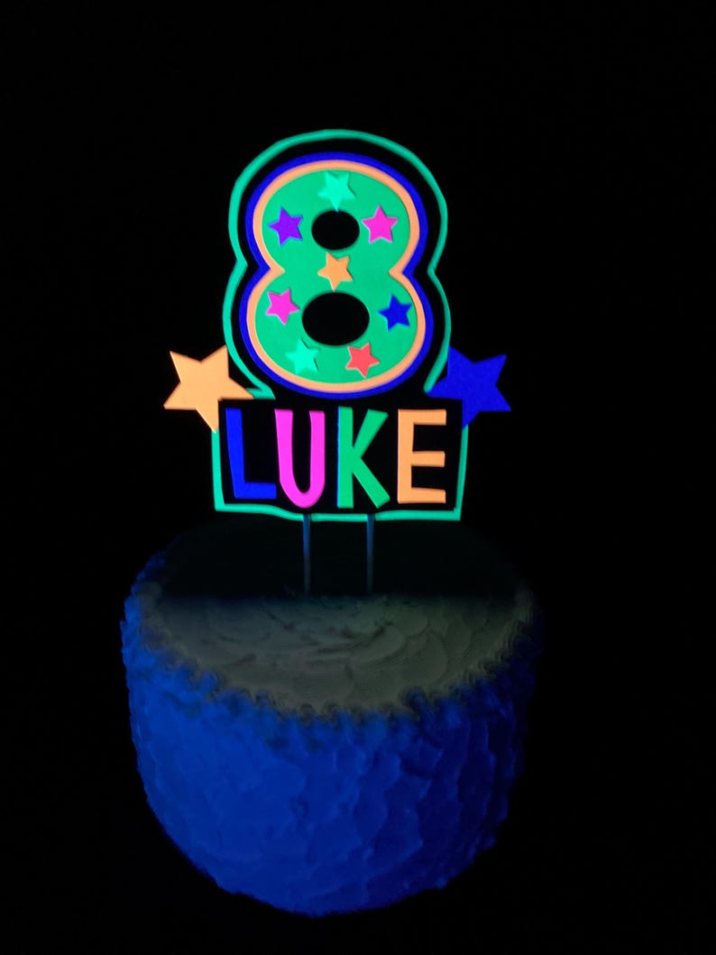 Puede incluir: Un adorno para pastel de color verde ne&oacute;n con el n&uacute;mero 8, estrellas de colores y el nombre "LUKE" en colores ne&oacute;n. El adorno para pastel est&aacute; sobre un pastel blanco glaseado.