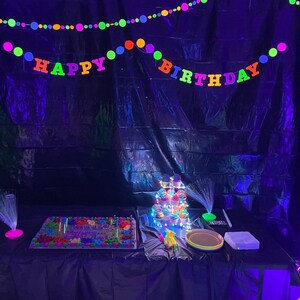 May include: A black backdrop with neon green, pink, orange, and yellow 'Happy Birthday' banner and a matching string of lights. A table with a black tablecloth has a neon green cake and a cupcake tower with neon green, pink, and blue frosting.