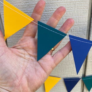 Peut inclure: Une guirlande color&eacute;e avec des drapeaux triangulaires jaunes, bleu sarcelle et bleus. Les drapeaux sont reli&eacute;s par un fil blanc. La guirlande est tenue dans une main sur un fond en bois clair.
