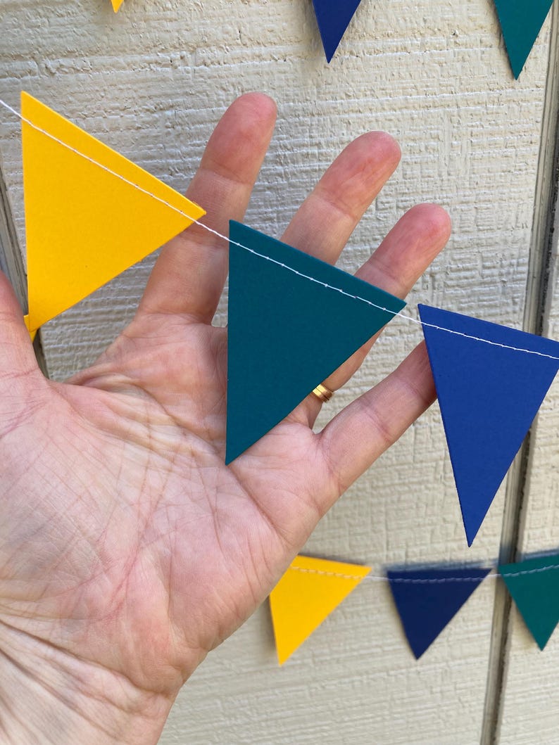 Peut inclure: Une guirlande color&eacute;e avec des drapeaux triangulaires jaunes, bleu sarcelle et bleus. Les drapeaux sont reli&eacute;s par un fil blanc. La guirlande est tenue dans une main sur un fond en bois clair.