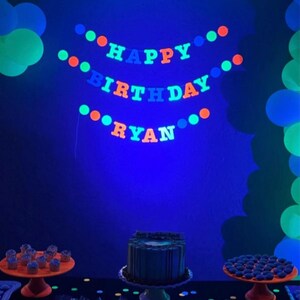 May include: A blue, green, and orange glow-in-the-dark banner that reads "Happy Birthday Ryan". The banner is hanging on a blue wall. There are two cakes on a table in front of the banner. One cake is a layered chocolate cake with white frosting and the other cake is a single-layer cake with blue frosting. There are also cupcakes on the table.